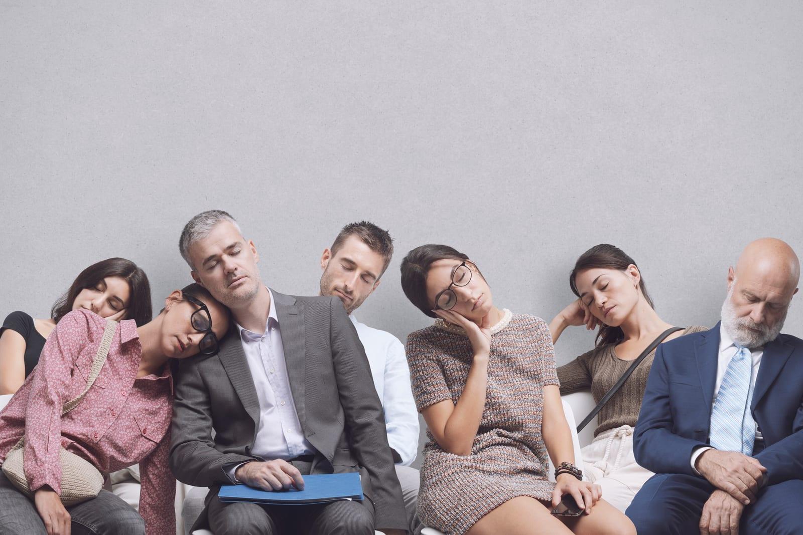 A row of eight people sitting in chairs against a plain grey wall, all with their eyes closed.